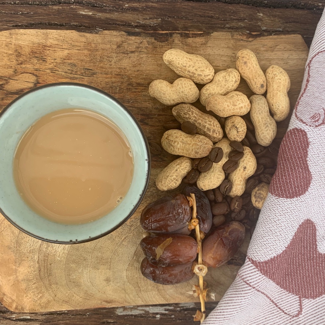 A cup of milk coffee with peanuts, dates and coffee beans nearby